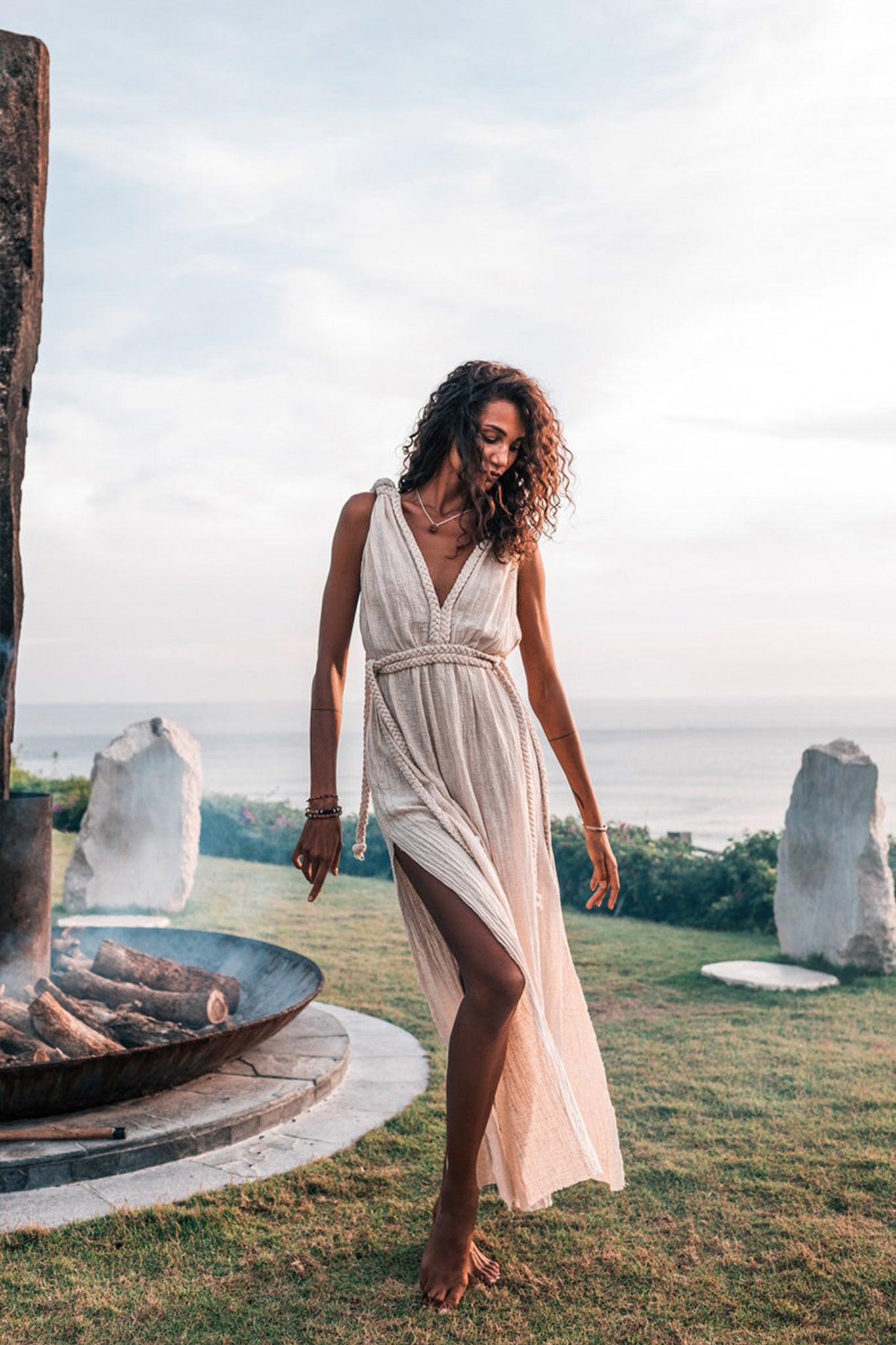 A woman with curly hair stands barefoot on the grass, showcasing the Off-White Nomad Spirit Dress by AYA Sacred Wear. This flowy boho dress features a deep V-neck and a slit, making it perfect for capturing picture-perfect moments near a large stone fire pit, set against the scenic ocean view and cloudy sky.