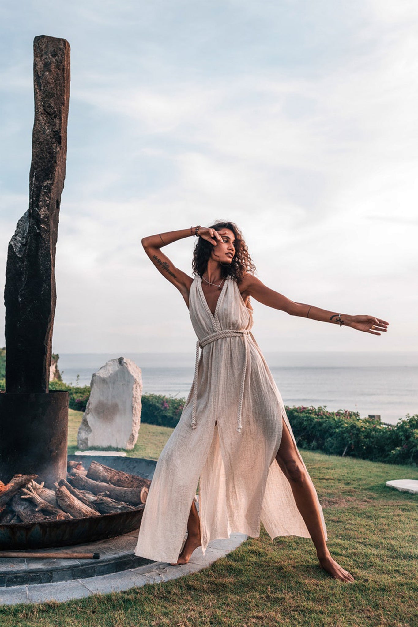 Wearing the Off-White Nomad Spirit Dress by AYA Sacred Wear, a woman dances gracefully beside a large campfire pit. She is outside on a grassy area overlooking the ocean beneath a cloudy sky.