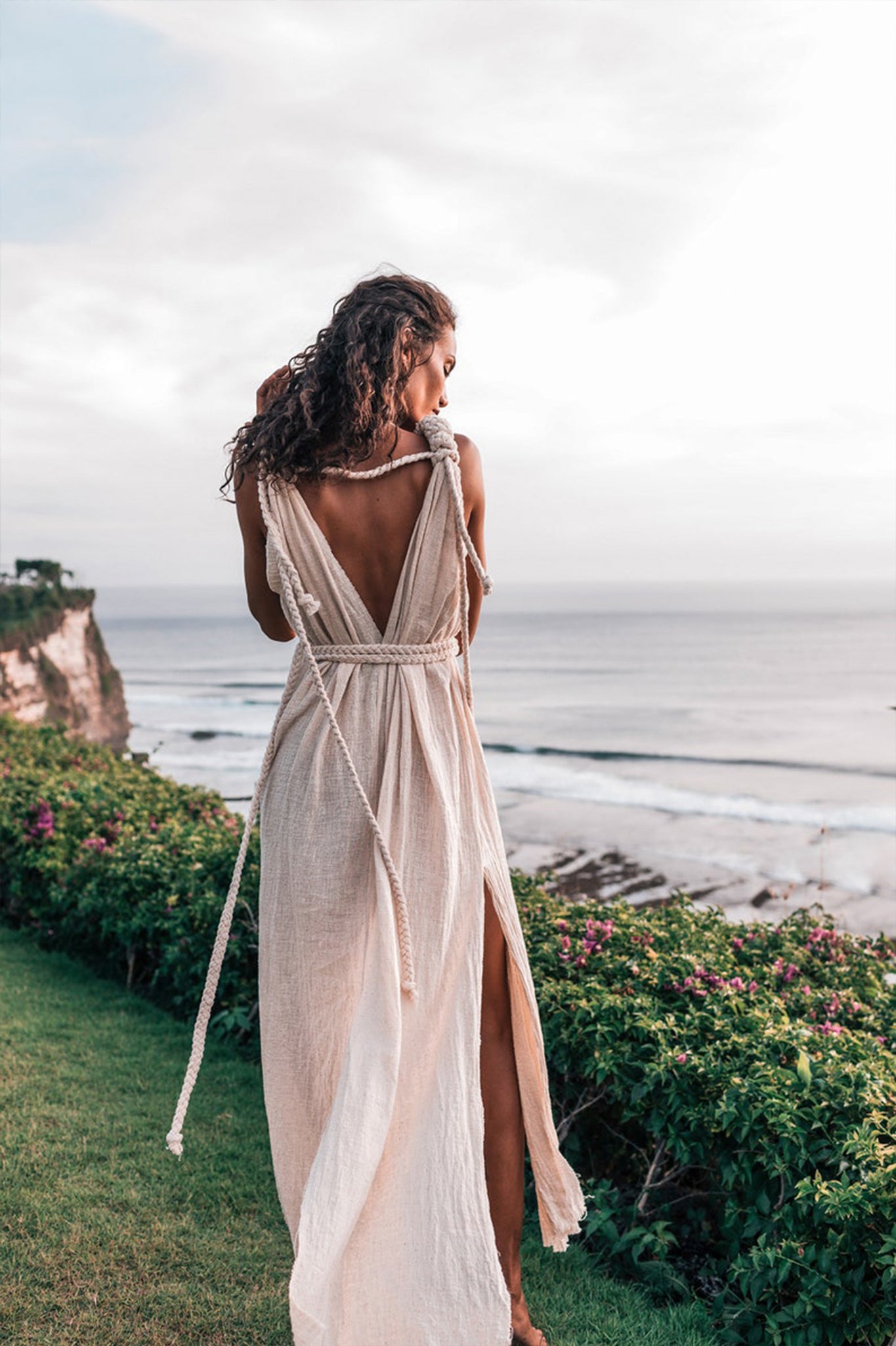 A woman stands gracefully on the grass, gazing at the ocean while wearing the Off-White Nomad Spirit Dress by AYA Sacred Wear. This flowing, boho-style dress, made from organic fabric, showcases a deep V-back and intricate tie details. Surrounded by lush greenery and a scenic coastal view, she embraces the tranquility of the cloudy sky.