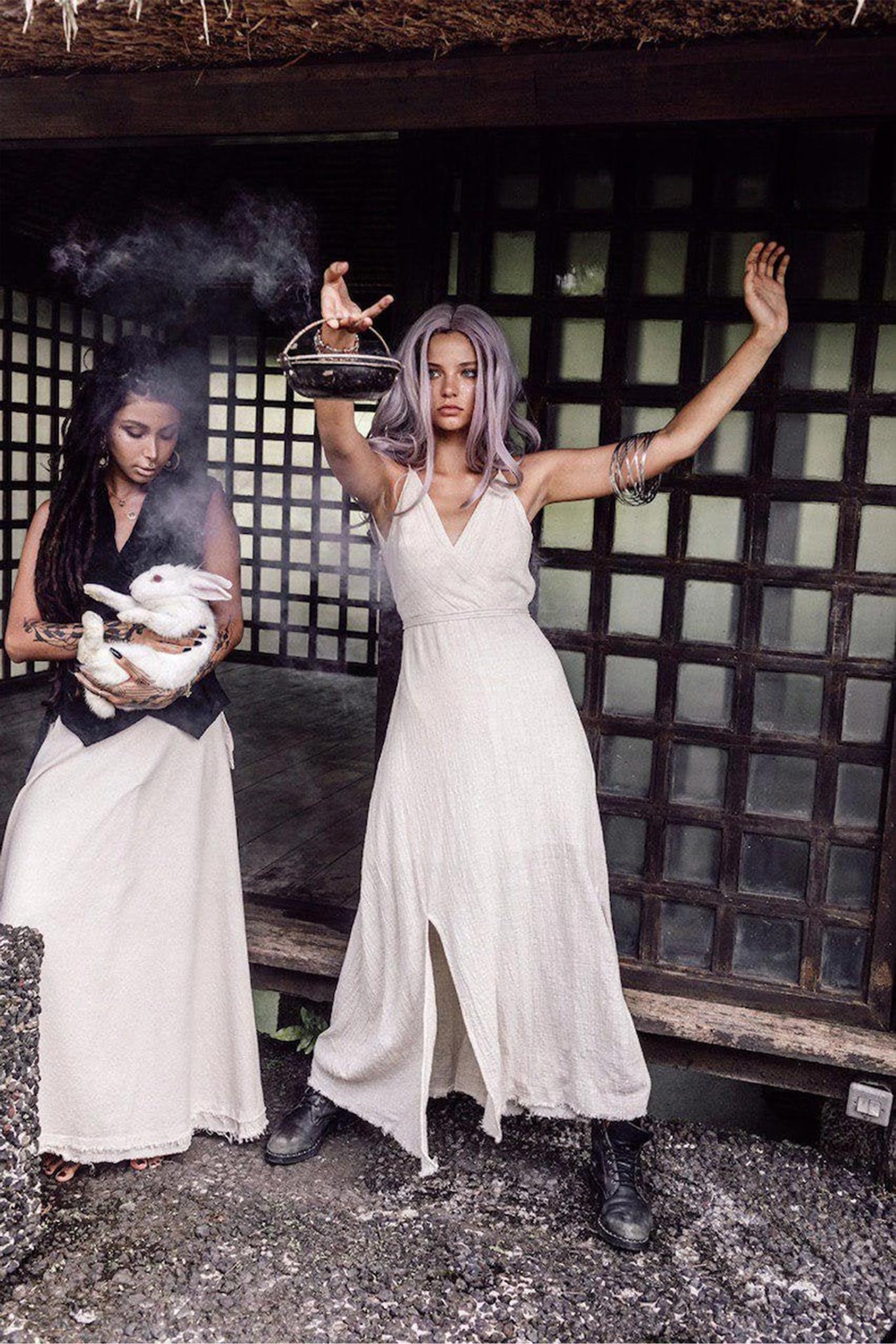 Two women in elegant off-white dresses from AYA Sacred Wear, reminiscent of a simple boho wedding style, stand outside a building with a grid window. One woman gently holds a white hen, while the other raises her hand with a smoking object. Their expressions are serious, and the rustic setting complements the open-back maxi dresses they wear.