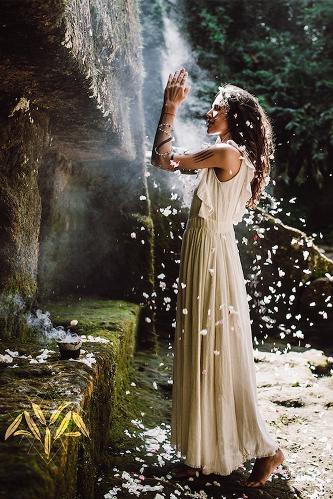 In the midst of a tranquil forest, a woman clad in an AYA Sacred Wear Simple Boho Bohemian Wedding Dress extends her hand toward sunlight streaming through the trees. Enveloped by flower petals, she is encompassed by a serene and mystical ambiance. Gentle smoke wafts from a small bowl placed on the mossy ground.