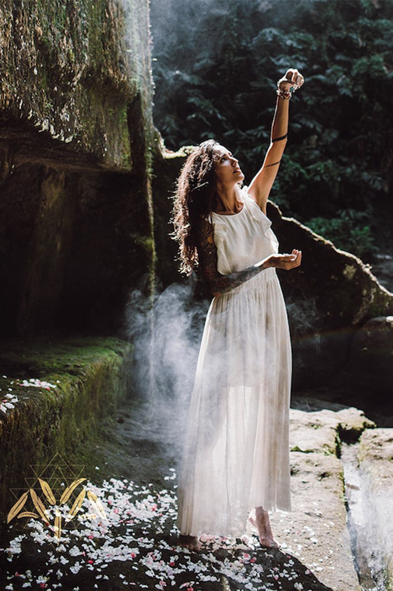 A woman, wearing the Simple Boho Bohemian Wedding Dress by AYA Sacred Wear, stands outdoors in a lush, green forest setting surrounded by rocky surfaces. She raises her hand towards the sky as mist envelops her and flower petals are scattered on the ground.