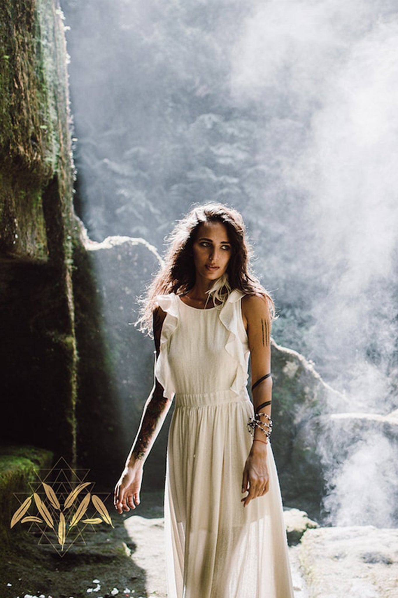 A woman in an elegant Simple Boho Bohemian Wedding Dress by AYA Sacred Wear stands in a misty, atmospheric setting, surrounded by mossy rocks and soft light filtering through.