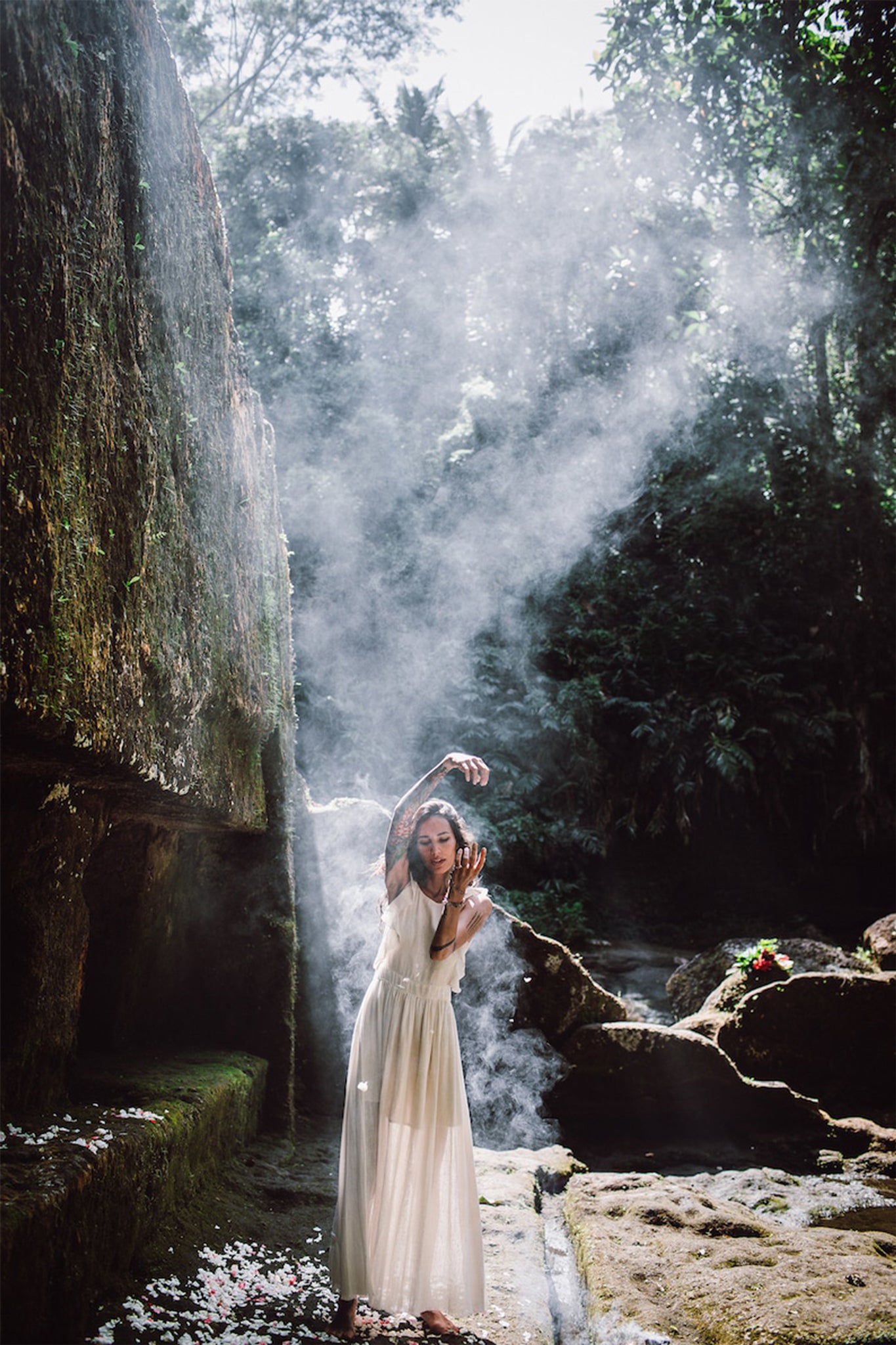 Draped in a Simple Boho Bohemian Wedding Dress by AYA Sacred Wear, a woman stands amidst a misty forest setting. She is surrounded by towering rocks and lush greenery, with sunlight filtering through to cast a soft glow around her. Her arms are gracefully raised, adding to the serene and ethereal atmosphere.