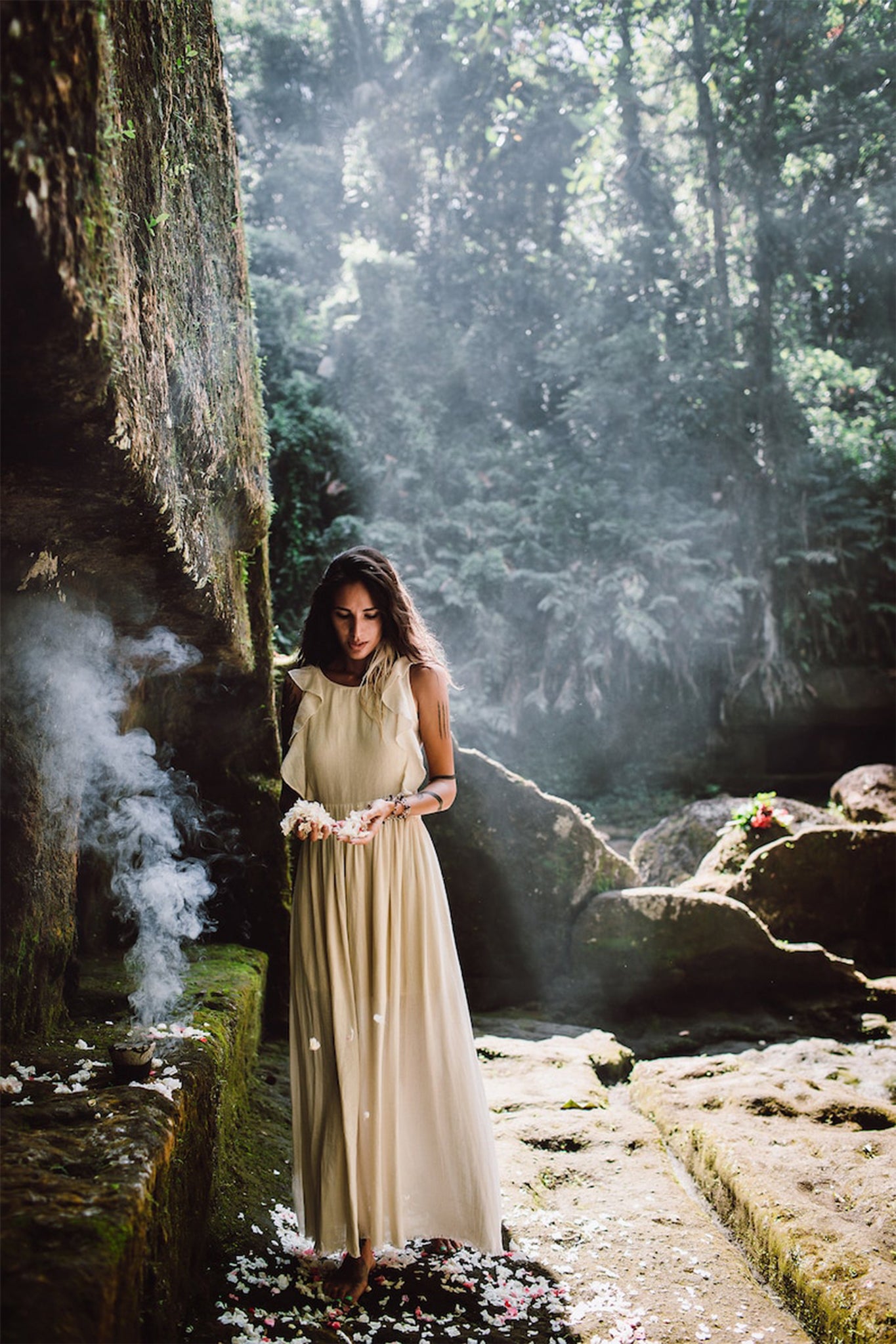 Dressed in a Simple Boho Bohemian Wedding Dress from AYA Sacred Wear, an individual stands in a tranquil forest setting, enveloped by mist and dappled sunlight. They are close to a rocky wall decorated with flowers, as fragrant smoke wafts upwards. Ferns and dense foliage provide a lush backdrop.