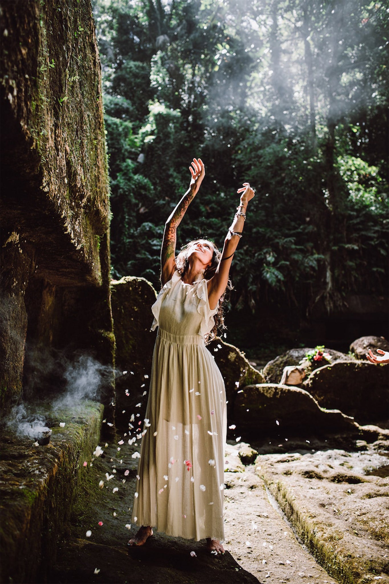 In a sunlit, lush forest clearing, a woman stands adorned in the Simple Boho Bohemian Wedding Dress by AYA Sacred Wear. Her arms are raised, and petals gracefully descend around her. Moss-covered rocks and gentle light filtering through the trees create an idyllic setting for a boho wedding moment.