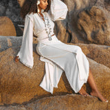 A woman with curly hair adorned with a white flower sits on a rock, wearing the Off-White Wrap Maxi Skirt from AYA Sacred Wear. This long, flowing skirt of organic handwoven cotton features intricate fringe, and she complements it with layered necklaces. The rocky background adds a natural, serene atmosphere to her boho style.