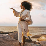 A woman in an elegant off-white wrap maxi skirt from AYA Sacred Wear stands barefoot on large rocks by the sea at sunset. Her curly hair dances in the wind as she holds a small flower, gazing thoughtfully towards the horizon. The sky is partly cloudy with warm tones, reflecting her serene expression.