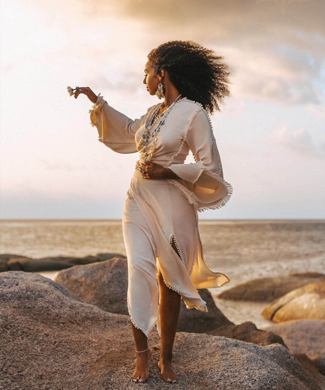 A woman in an elegant off-white wrap maxi skirt from AYA Sacred Wear stands barefoot on large rocks by the sea at sunset. Her curly hair dances in the wind as she holds a small flower, gazing thoughtfully towards the horizon. The sky is partly cloudy with warm tones, reflecting her serene expression.