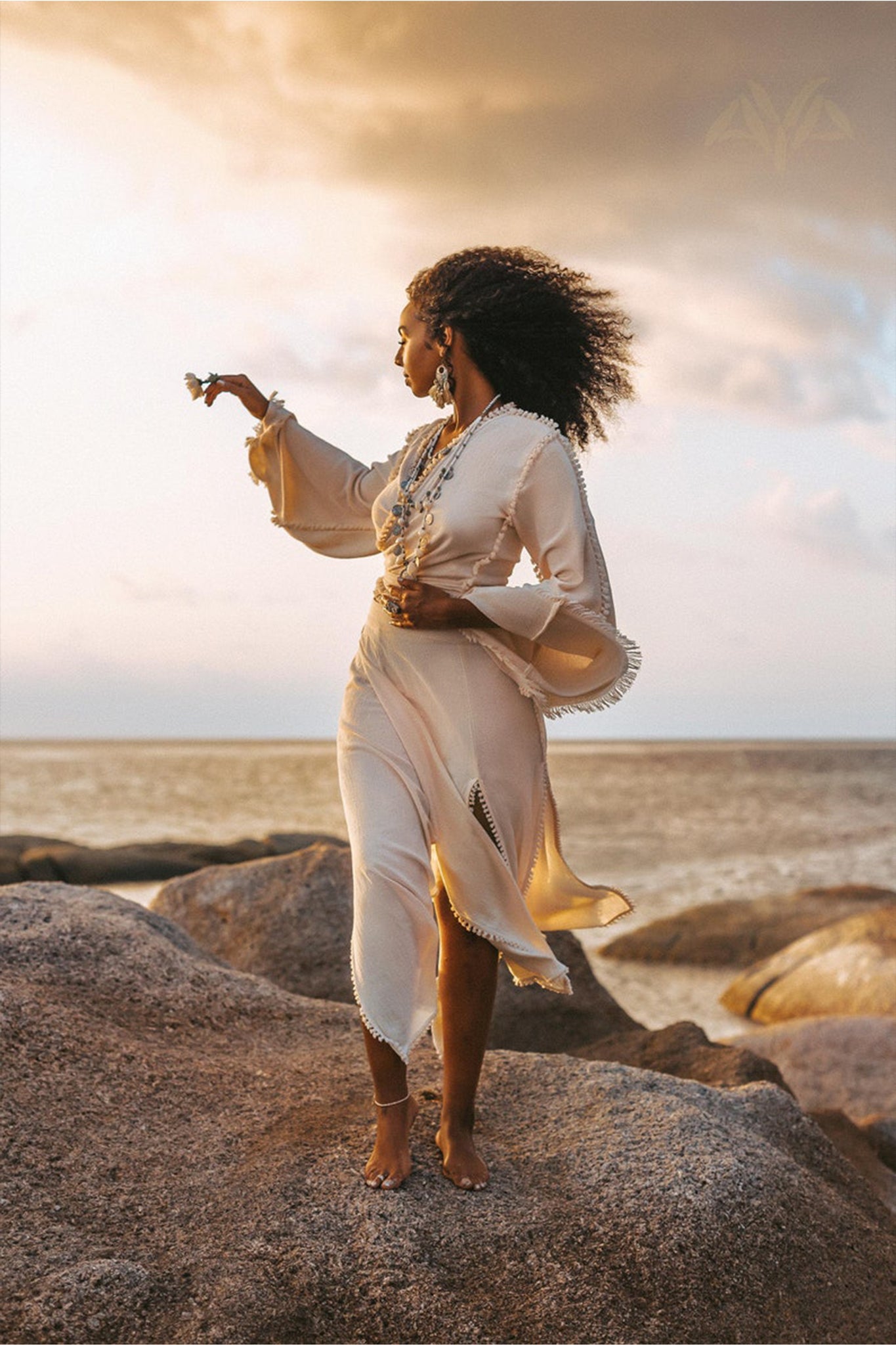 A woman in an elegant off-white wrap maxi skirt from AYA Sacred Wear stands barefoot on large rocks by the sea at sunset. Her curly hair dances in the wind as she holds a small flower, gazing thoughtfully towards the horizon. The sky is partly cloudy with warm tones, reflecting her serene expression.