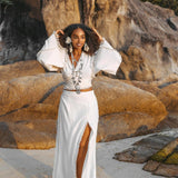A woman wearing an Off-White Wrap Maxi Skirt by AYA Sacred Wear strolls along a sandy beach, her outfit beautifully complemented by jewelry. Amidst large rocks and lush greenery, the sunlight highlights the intricate handmade craftsmanship of her boho-style long cotton skirt.