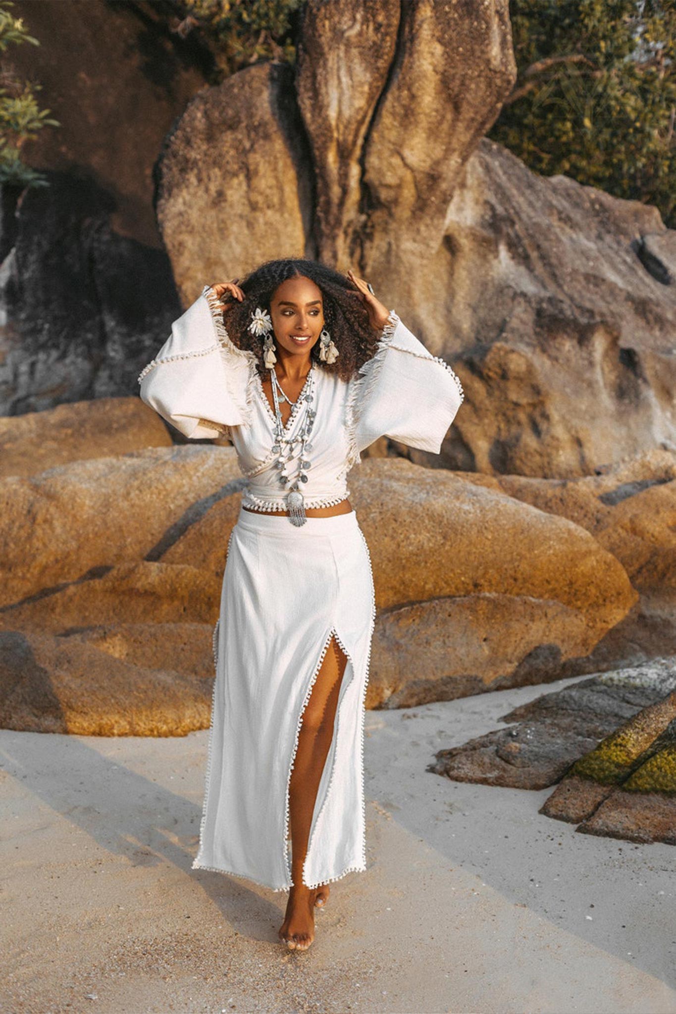 A woman wearing an Off-White Wrap Maxi Skirt by AYA Sacred Wear strolls along a sandy beach, her outfit beautifully complemented by jewelry. Amidst large rocks and lush greenery, the sunlight highlights the intricate handmade craftsmanship of her boho-style long cotton skirt.