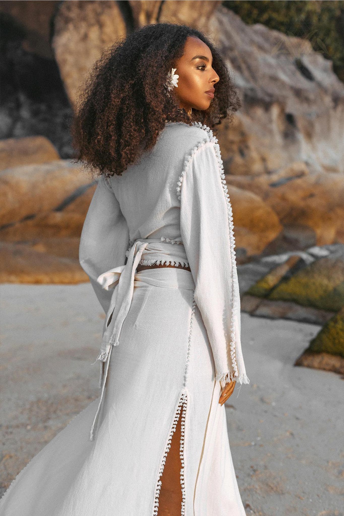 An elegant woman with curly hair stands on a sandy beach, wearing the Off-White Wrap Maxi Skirt from AYA Sacred Wear. Crafted as a boho long skirt from organic handwoven cotton, the look is completed with a white flower in her hair. She gazes to the side, framed by large rocks and moss in the background.