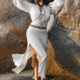 Barefoot on rocks, a person wears the Off-White Wrap Maxi Skirt from AYA Sacred Wear, a flowing boho creation embellished with pearl accents and handmade knots on organic handwoven cotton. Adorned in layered jewelry and large earrings, they place a hand on their head and gaze into the distance under natural sunlight.