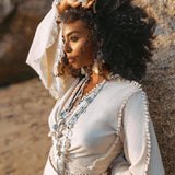 A woman with curly hair is on the beach, dressed in an AYA Sacred Wear ensemble featuring a long-sleeved outfit with fringe details and an Off-White Wrap Maxi Skirt - a handmade boho long cotton skirt. She has a flower tucked in her hair and layered necklaces. The warm, golden sunlight bathes the scene, accentuating the serene texture of her organic handwoven cotton attire.
