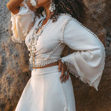 Dressed in a textured off-white ensemble, a woman poses outdoors against a rocky backdrop. Shielding her eyes with one hand, she accessorizes with large earrings and layered necklaces. Her outfit includes bell sleeves made from organic handwoven cotton and an Off-White Wrap Maxi Skirt by AYA Sacred Wear, featuring a boho style with slits.