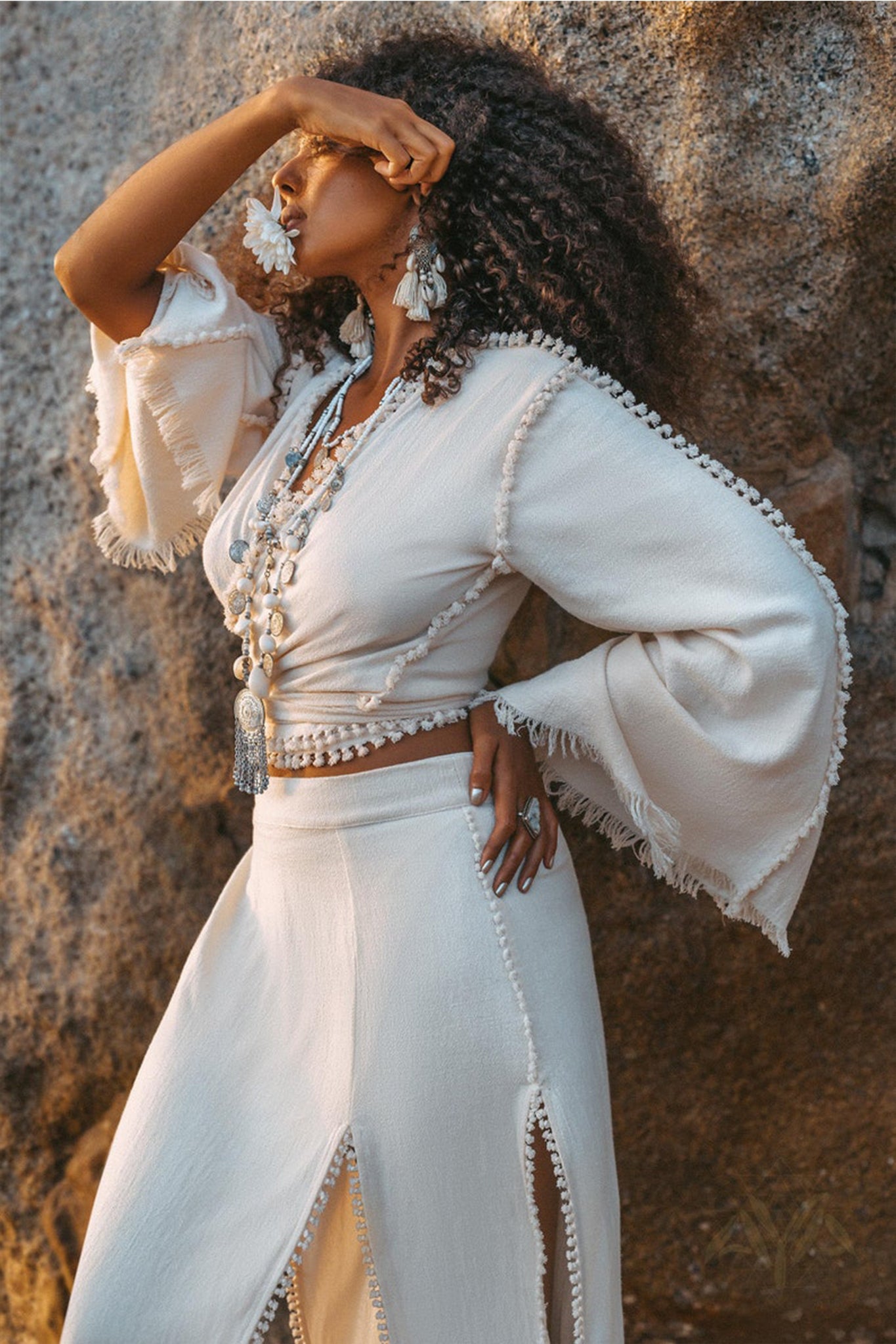 Dressed in a textured off-white ensemble, a woman poses outdoors against a rocky backdrop. Shielding her eyes with one hand, she accessorizes with large earrings and layered necklaces. Her outfit includes bell sleeves made from organic handwoven cotton and an Off-White Wrap Maxi Skirt by AYA Sacred Wear, featuring a boho style with slits.