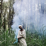 A person dressed in an Off-White Long Boho Skirt with Pockets from AYA Sacred Wear and a headscarf stands amidst tall green grass and trees. A light haze of smoke or mist creates a mystical ambiance in the forest setting. The person appears to be in a serene and contemplative state.