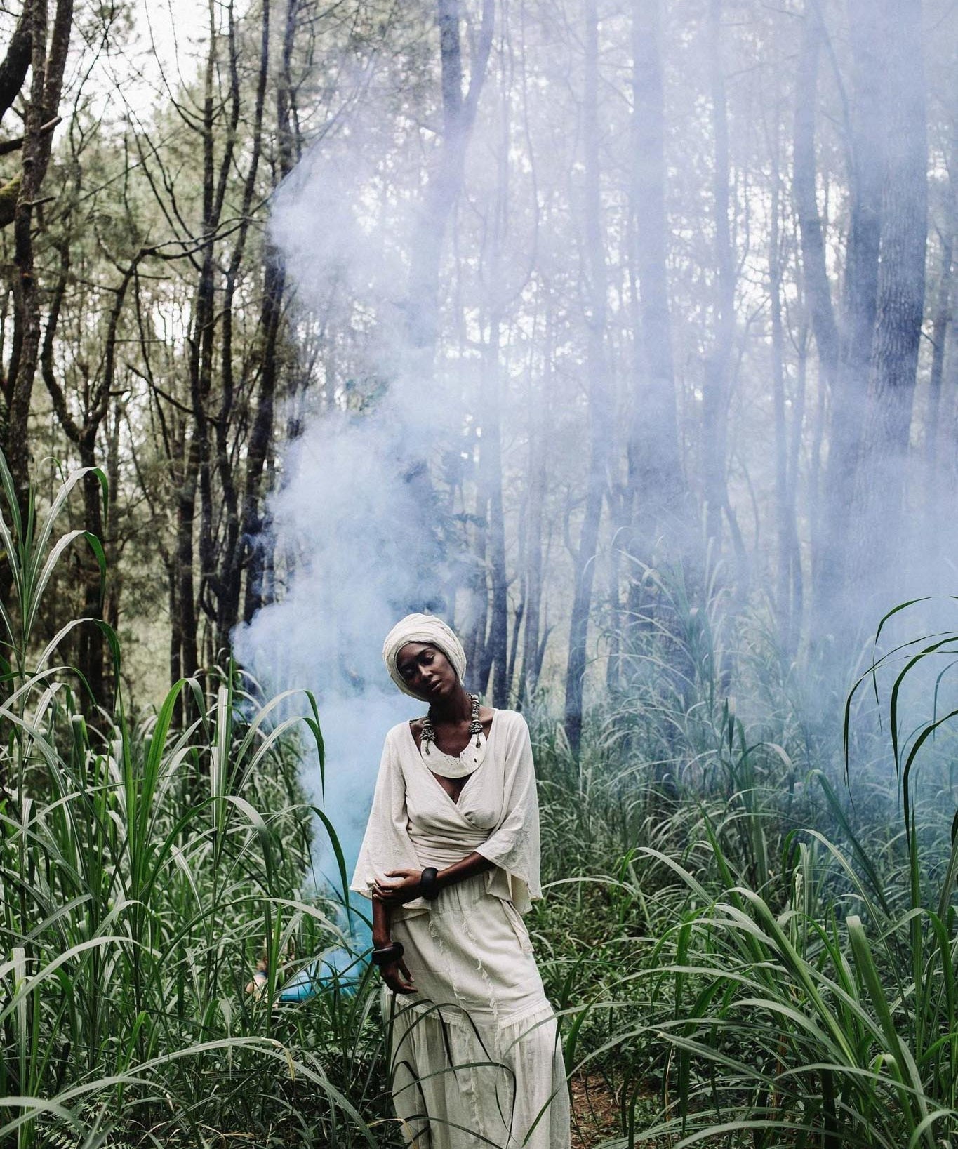 A person dressed in an Off-White Long Boho Skirt with Pockets from AYA Sacred Wear and a headscarf stands amidst tall green grass and trees. A light haze of smoke or mist creates a mystical ambiance in the forest setting. The person appears to be in a serene and contemplative state.