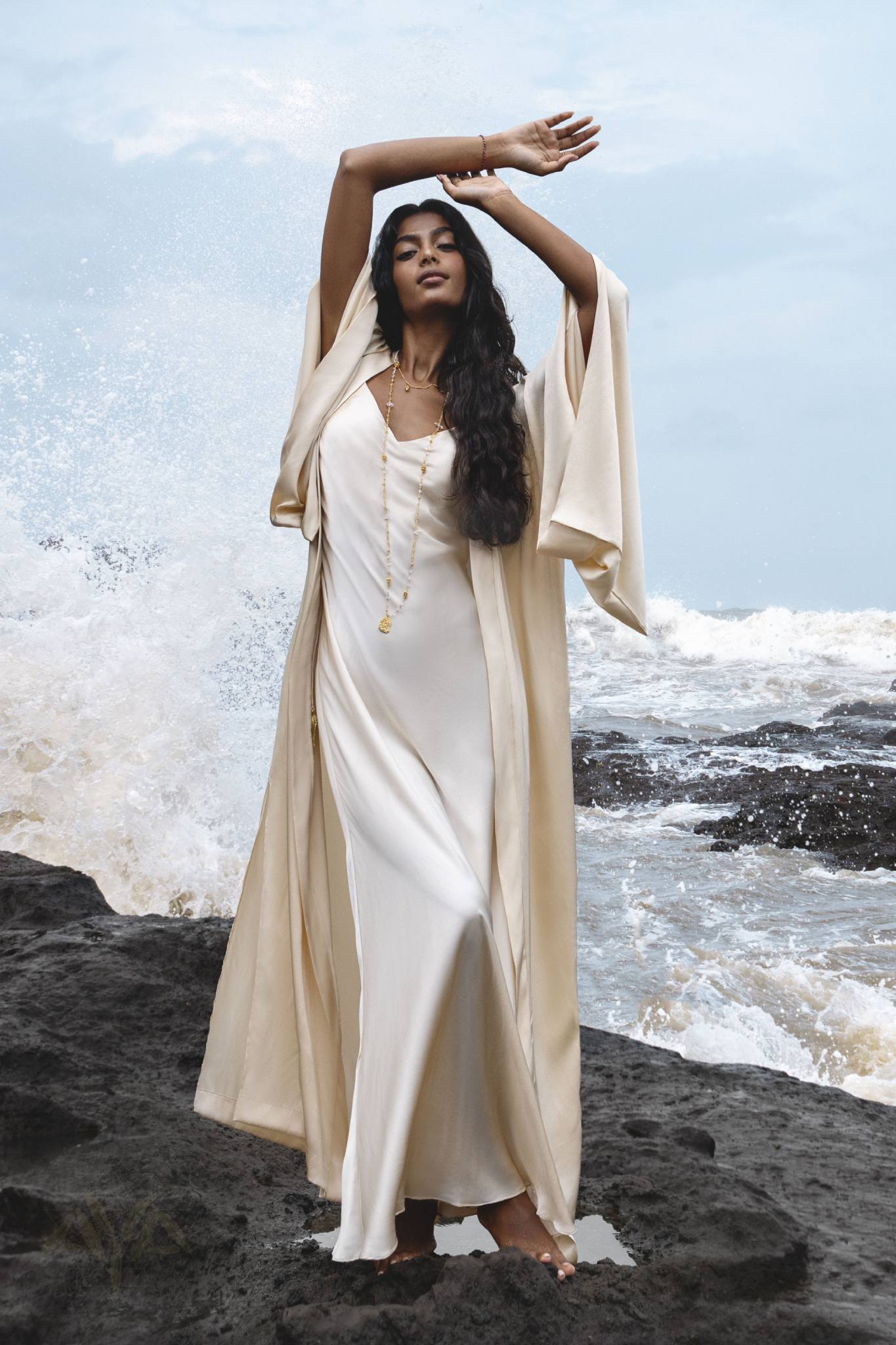 A person gracefully poses on a rocky shoreline, wearing the AYA Sacred Wear Cream Silk Kimono Cover Up with a long necklace. Waves splash dramatically in the background against an elegantly overcast sky.