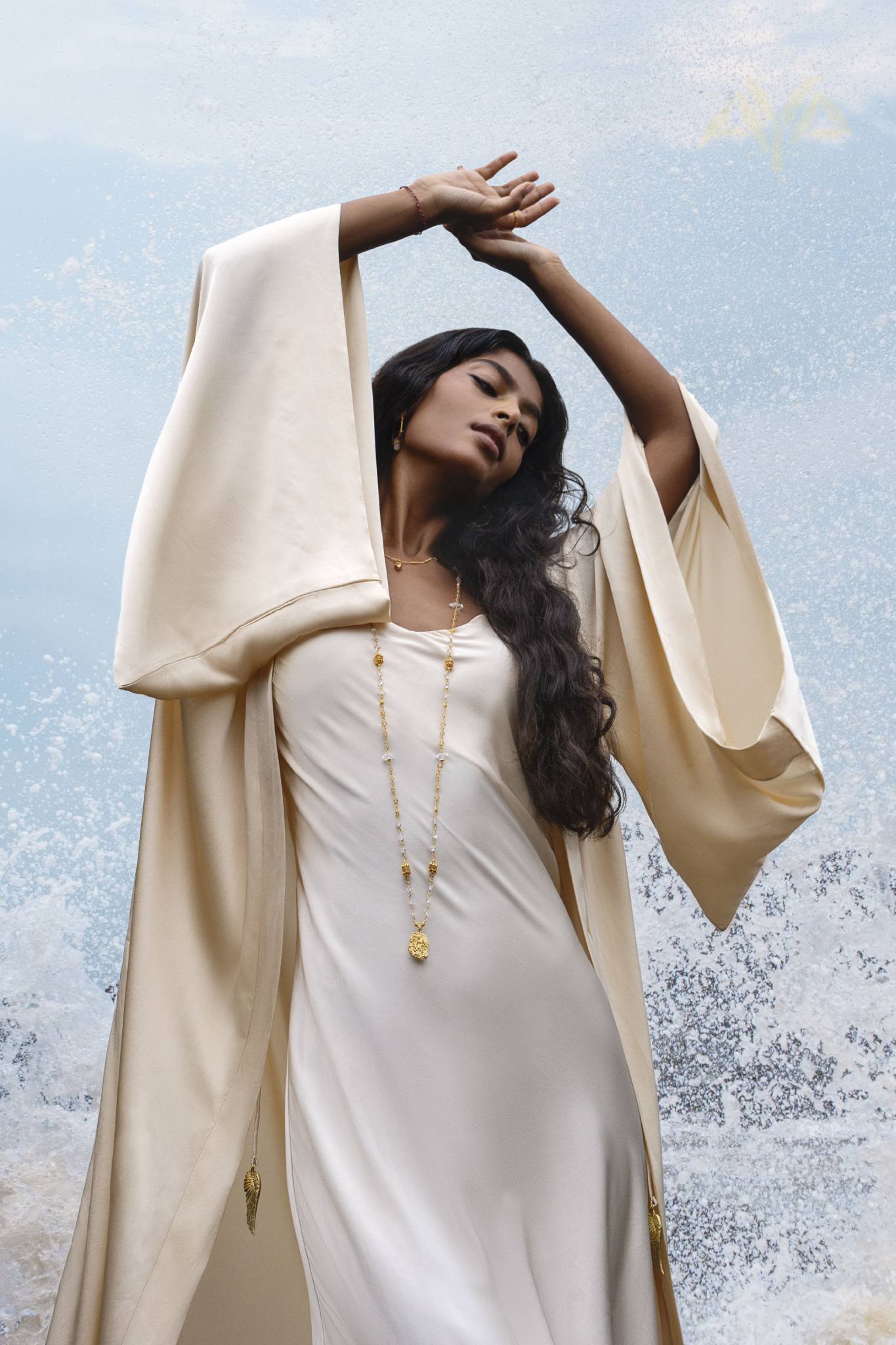 A woman in a Cream Silk Wide Sleeve Bohemian Cardigan Up by AYA Sacred Wear stands with her eyes closed, arms raised. Her dark hair cascades over a long necklace, and her attire enhances the textured blue and white background reminiscent of sky and sea.