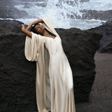 Someone in an elegant Cream Silk Kimono Cover Up by AYA Sacred Wear leans back gracefully against a large rock by the sea, as waves crash dramatically behind, creating a tranquil scene that evokes a serene connection with nature.