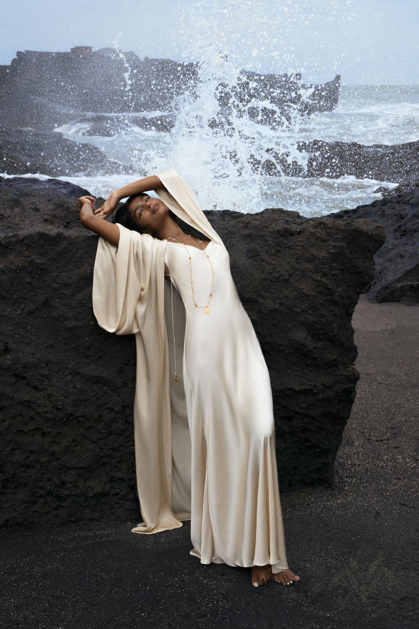 Someone in an elegant Cream Silk Kimono Cover Up by AYA Sacred Wear leans back gracefully against a large rock by the sea, as waves crash dramatically behind, creating a tranquil scene that evokes a serene connection with nature.