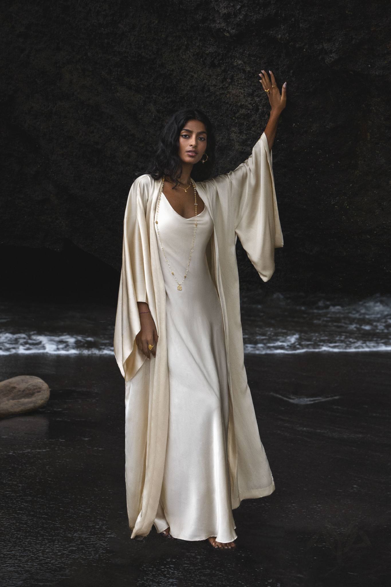 A woman poses gracefully by dark rocks near the water in a Cream Ahimsa Silk Kimono Cardigan by AYA Sacred Wear. Her serene expression complements her long, wavy hair and elegant necklace, embodying sustainable living with this wide-sleeve Bohemian cardigan. - make an etsy alt text  use one Wide Sleeve Caftan • Kimono Dress Women key words