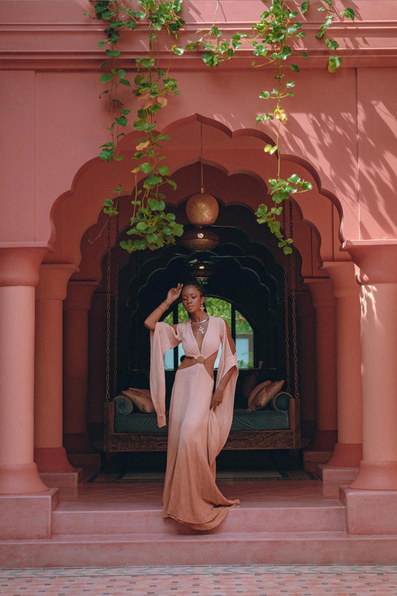 Dressed in an Ombré Dusty Pink Botanically Dyed Dress—an open-belly maxi bridesmaid piece by AYA Sacred Wear—a person stands gracefully in an ornate, arched corridor decorated with lush hanging greenery. A plush bench is situated behind them, enhancing the tranquil and opulent ambience.