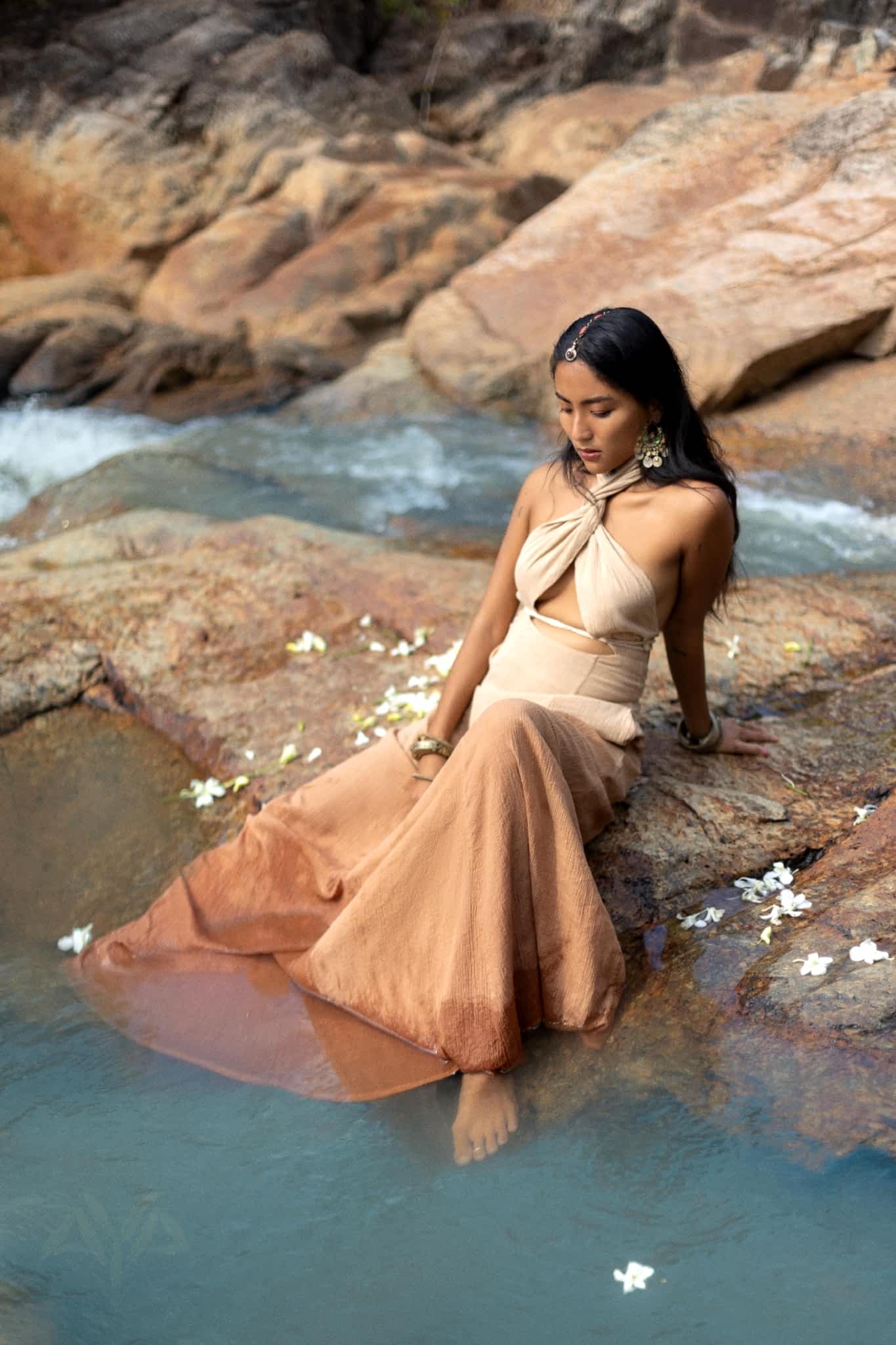 A woman wearing the AYA Sacred Wear Aphrodite Dress, an Ombre Powder Ohra Harmonia and Boho Greek Goddess style dress, sits on a rock by a flowing river with scattered white flowers around her and her feet dipped in clear water amidst natural rock formations.