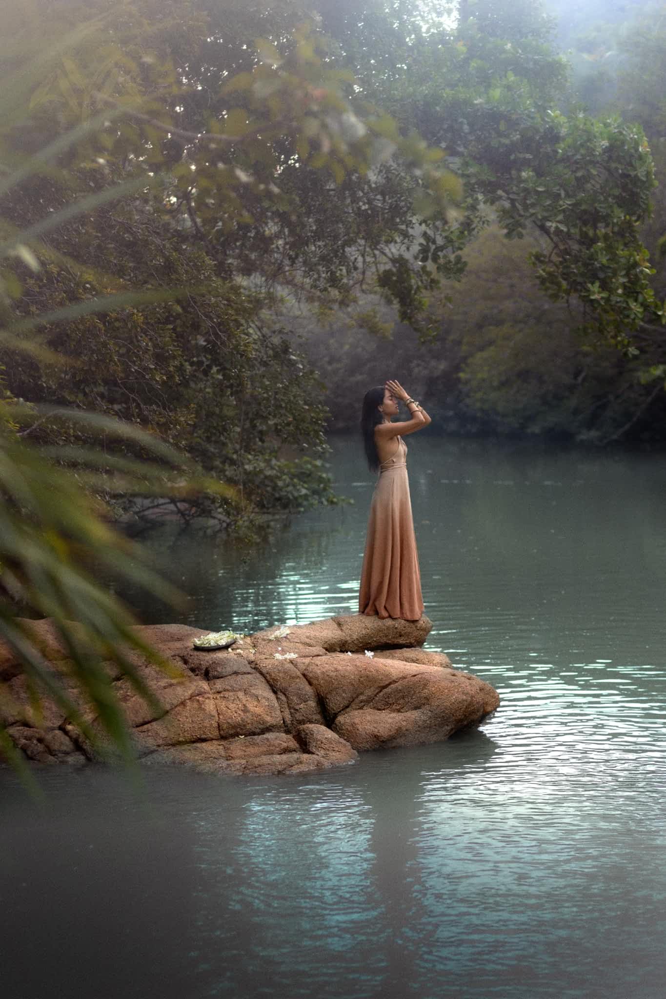 A woman in an Aphrodite Dress from AYA Sacred Wear, made with handwoven fabric and botanical dyes, stands on rocks by a serene river. She gazes upwards, surrounded by lush trees and mist, creating a tranquil and ethereal atmosphere.