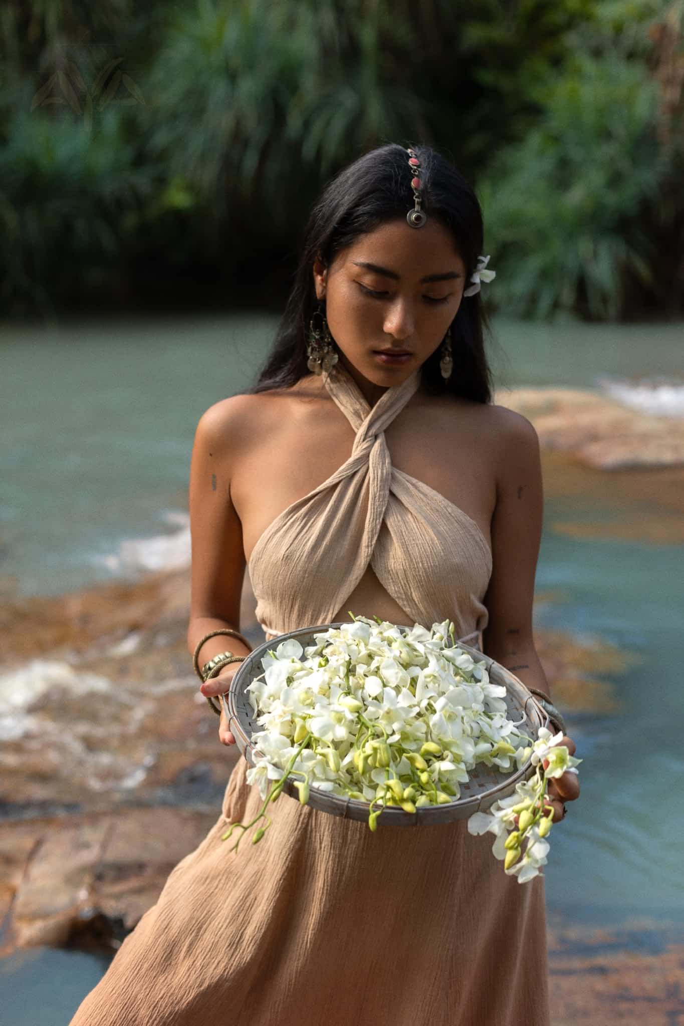 A person with long dark hair stands by a serene body of water, holding a large bowl of white flowers. They wear an Aphrodite Dress from AYA Sacred Wear, featuring handwoven fabric in a beige halter style adorned with criss-cross details and have a blue flower behind their ear. The lush greenery surrounds them.