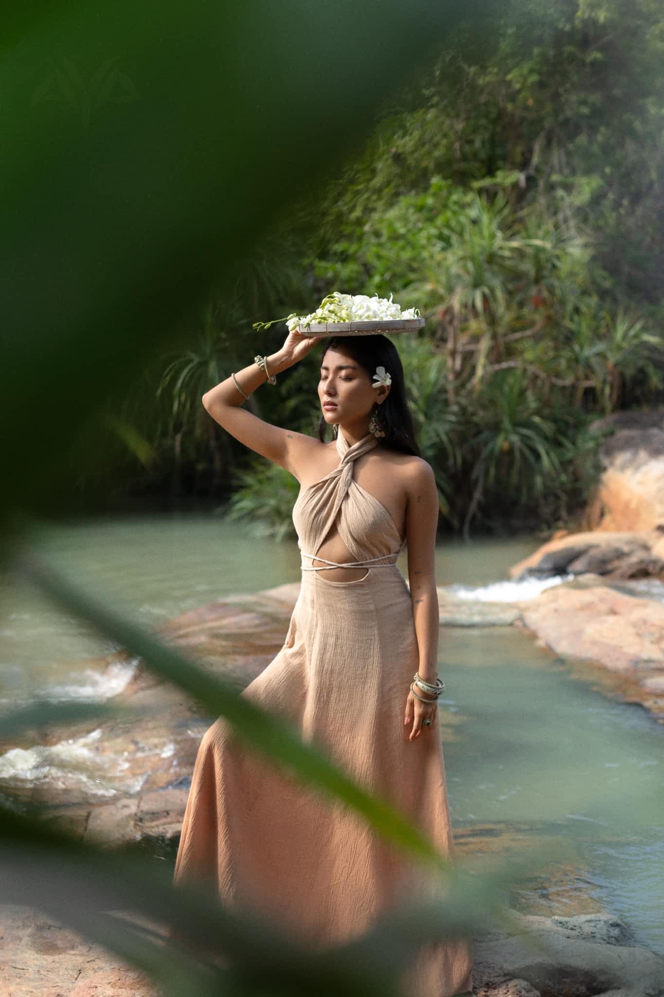 A woman in an Aphrodite Dress by AYA Sacred Wear, crafted from handwoven fabric, stands by a riverbank with a flower tray. She gazes thoughtfully into the lush greenery, as a large leaf partly obstructs the foreground view.