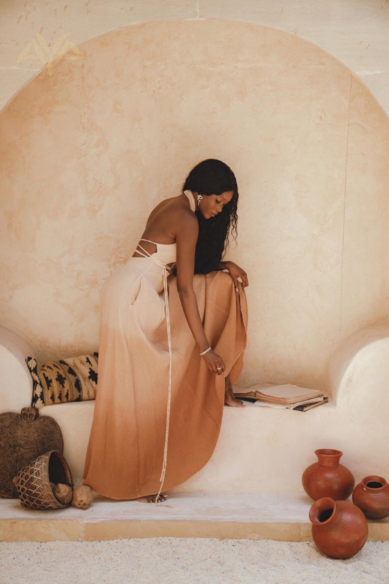 A woman in an Ombre Powder Ohra Harmonia Dress by AYA Sacred Wear leans in a stone alcove, surrounded by decorative vases and pillows. Crafted from handwoven fabric with botanical dyes, her attire complements her wavy hair. She is adorned with earrings and bracelets, with an open book beside her.