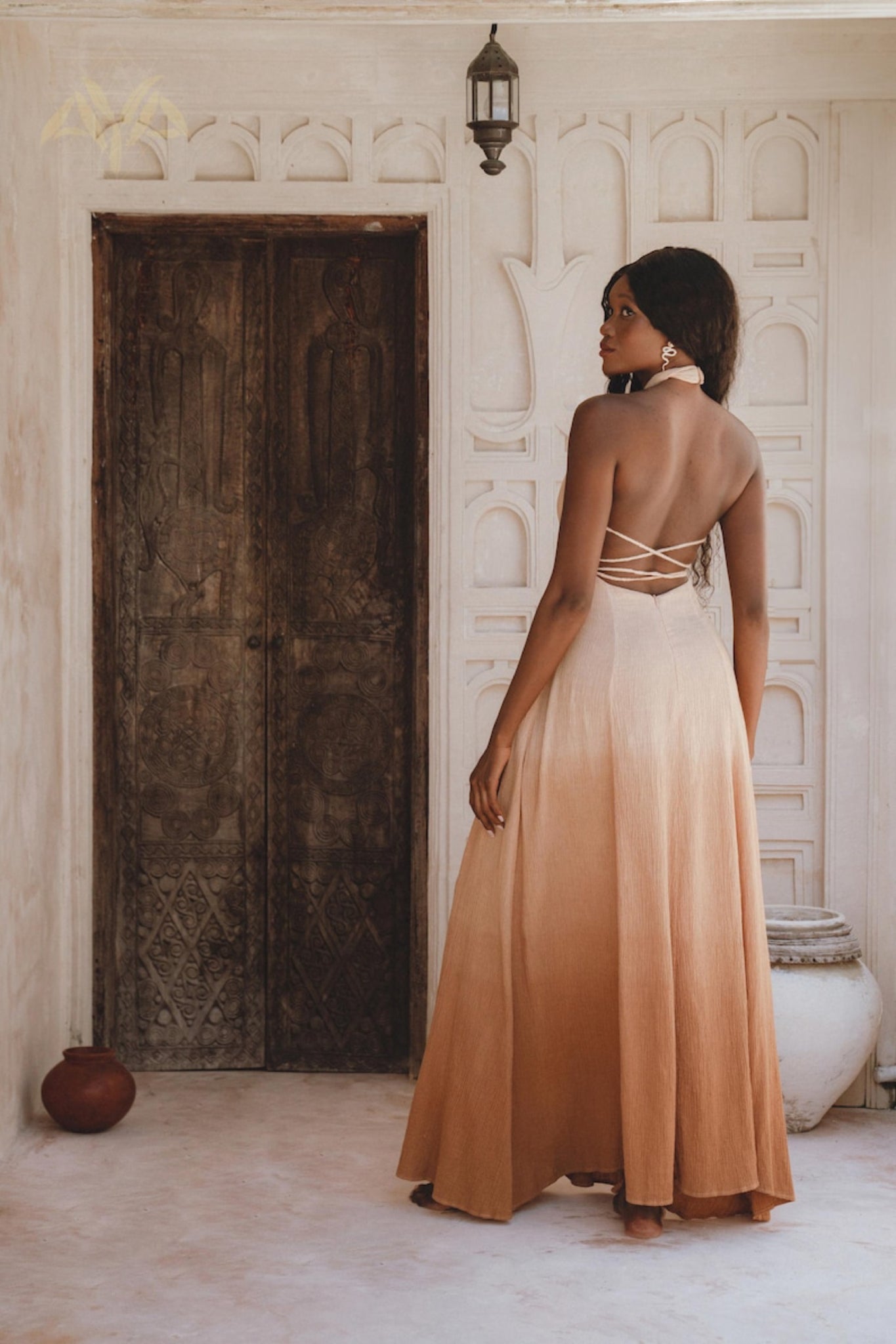 In a rustic room, a woman wears the AYA Sacred Wear Ombre Powder Ohra Harmonia Dress in handwoven fabric. Facing a carved wooden door with her back to the camera, the scene is enriched by decorative pots and lanterns on white textured walls, enhanced with botanical dyes.