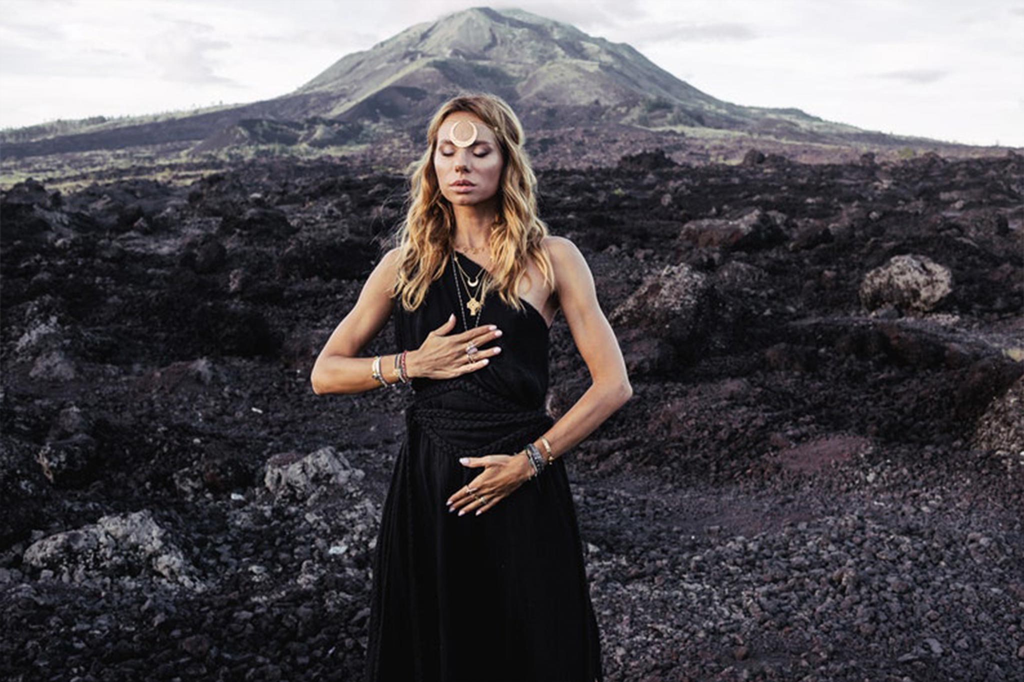 A woman stands in a rocky, volcanic landscape with a mountain in the background. She wears an Organic Black Boho One Shoulder Dress by AYA Sacred Wear, adorned with jewelry. With her eyes closed, one hand on her chest and the other on her abdomen, she exudes a sense of tranquility.