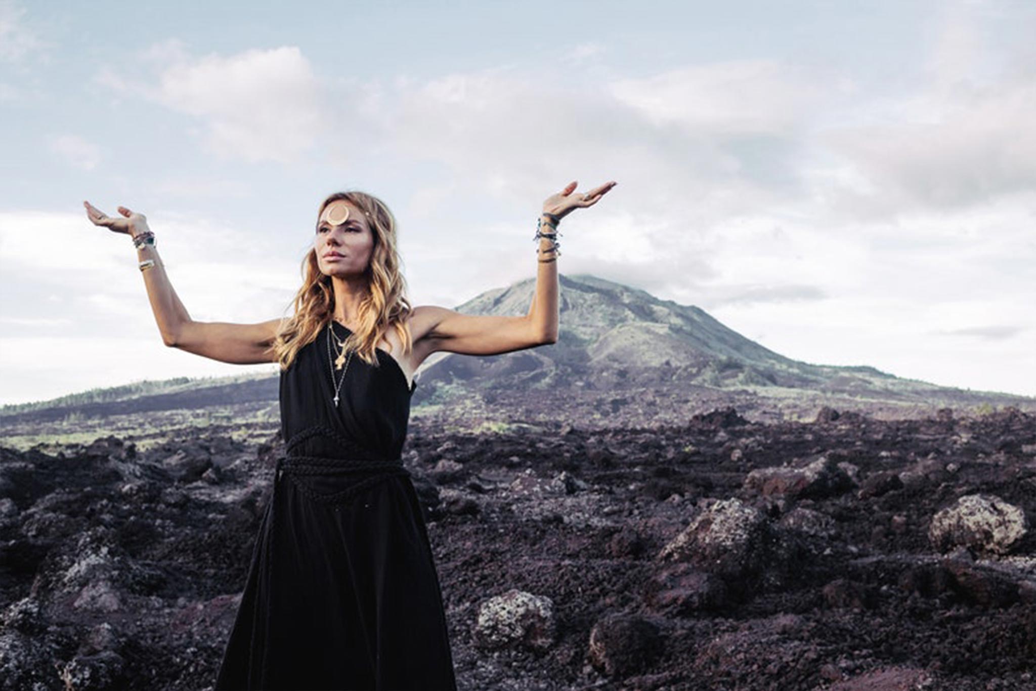 Wearing the Organic Black Boho One Shoulder Dress from AYA Sacred Wear, a woman stands on rocky terrain with her arms raised against a backdrop of mountains and a cloudy sky. Her serene expression is highlighted by bracelets and a headband, perfectly complementing the dress's organic fabric as it gently flows around her in harmony with the natural surroundings.