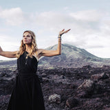 Wearing the Organic Black Boho One Shoulder Dress from AYA Sacred Wear, a woman stands on rocky terrain with her arms raised against a backdrop of mountains and a cloudy sky. Her serene expression is highlighted by bracelets and a headband, perfectly complementing the dress's organic fabric as it gently flows around her in harmony with the natural surroundings.