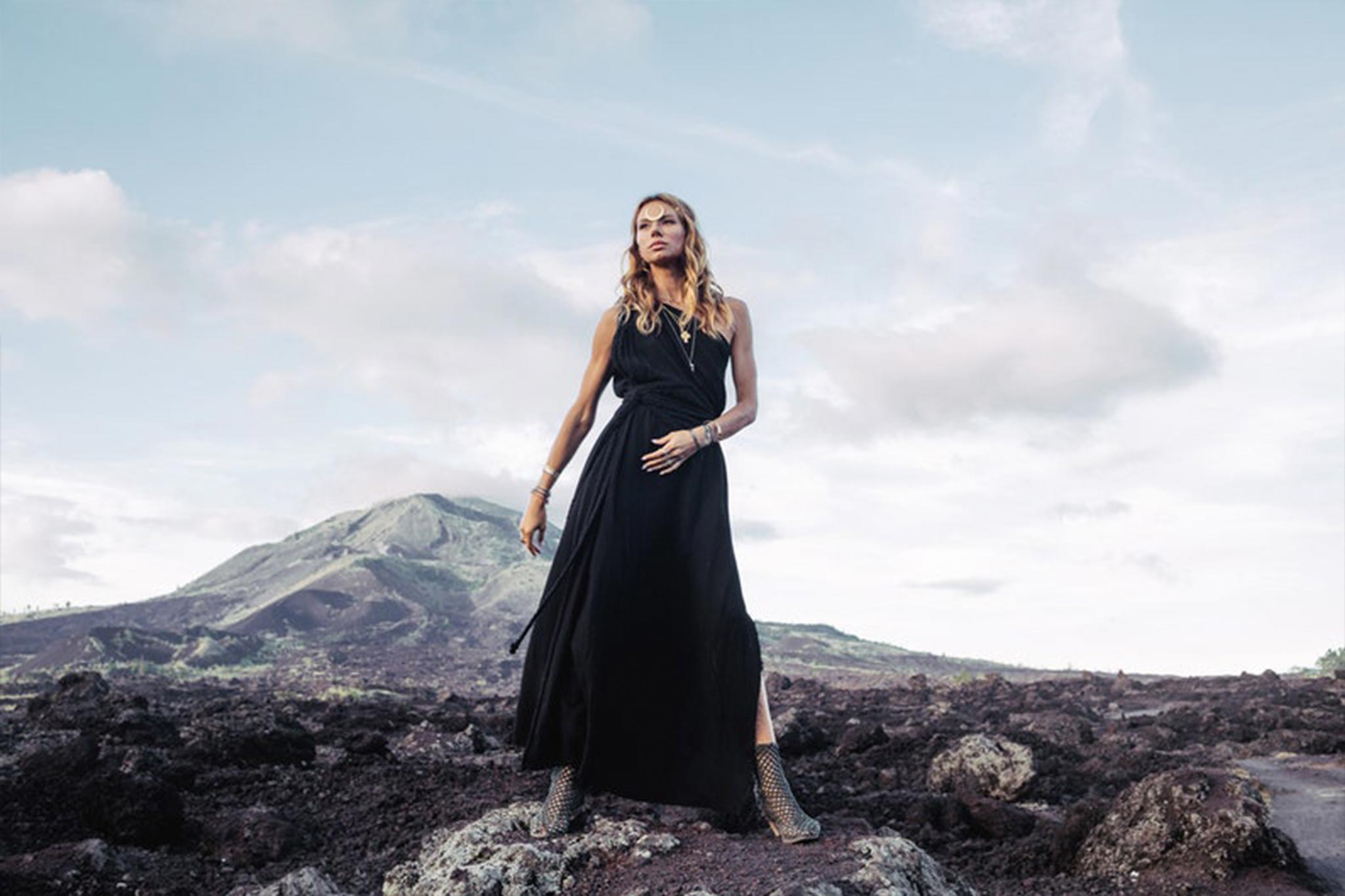 Clad in the Organic Black Boho One Shoulder Dress by AYA Sacred Wear, a person stands confidently on rocky terrain, framed by a mountainous landscape in the background under a partly cloudy sky.