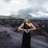 A person in an Organic Black Boho One Shoulder Dress by AYA Sacred Wear stands on a rocky landscape with a volcanic mountain in the background. They form a heart shape with their hands at their waist, highlighted by a braided belt. The sky above is cloudy.