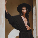 A model poses elegantly in an oversized, hand-knotted black jumpsuit featuring asymmetrical design elements. She stands against a textured wall, wearing a wide-brimmed black hat and layered jewelry, highlighting the stylish and sophisticated features of the jumpsuit.