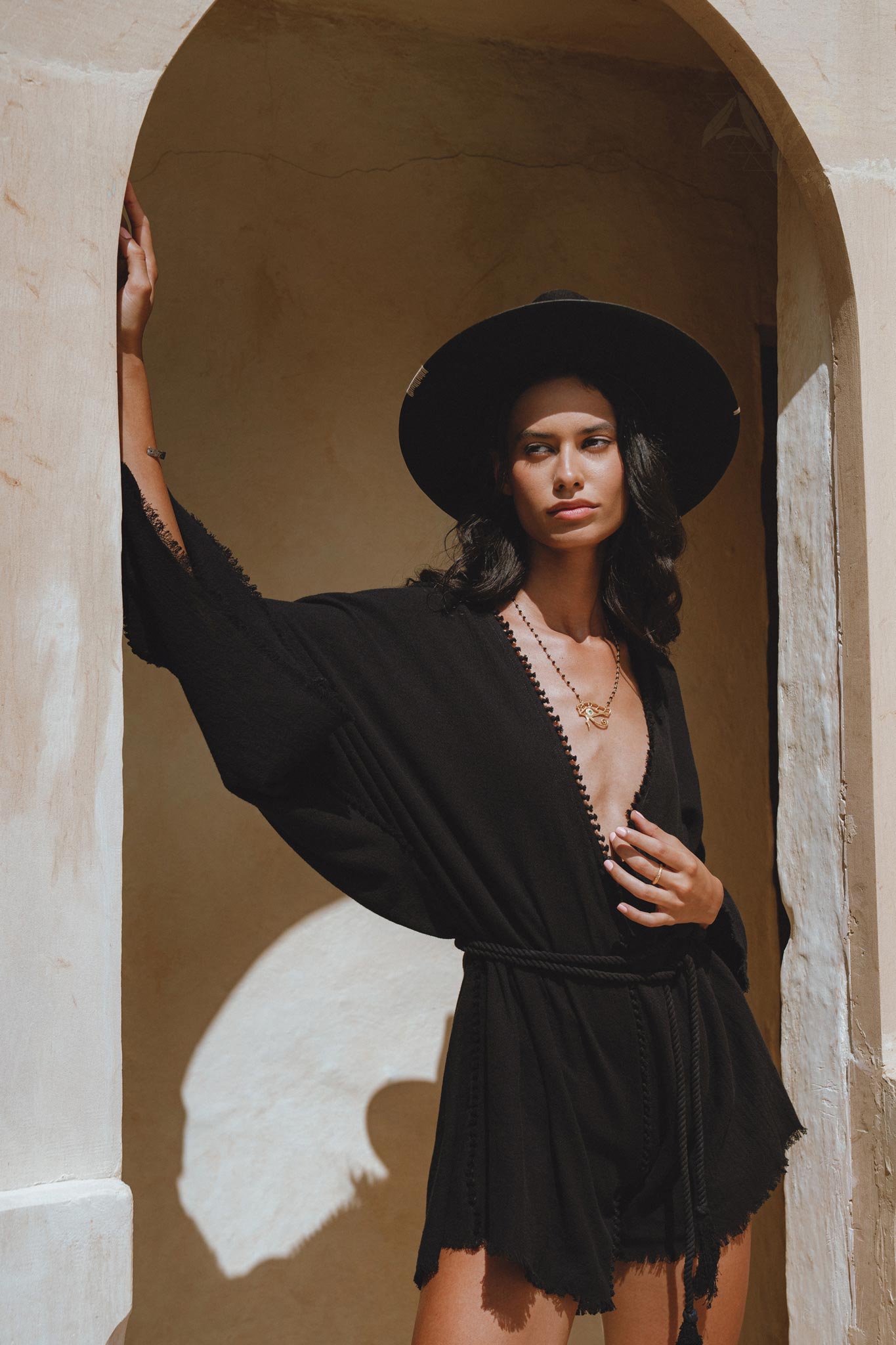 A model poses elegantly in an oversized, hand-knotted black jumpsuit featuring asymmetrical design elements. She stands against a textured wall, wearing a wide-brimmed black hat and layered jewelry, highlighting the stylish and sophisticated features of the jumpsuit.