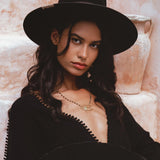 A woman poses confidently in an oversized hand-knotted black jumpsuit, featuring an elegant and asymmetrical design. She wears a stylish black hat and a delicate necklace, set against a warm, textured background that enhances the sophisticated look of the outfit. Perfect for showcasing the contemporary fashion aesthetic of the Oversized Hand Knotted Jumpsuit.