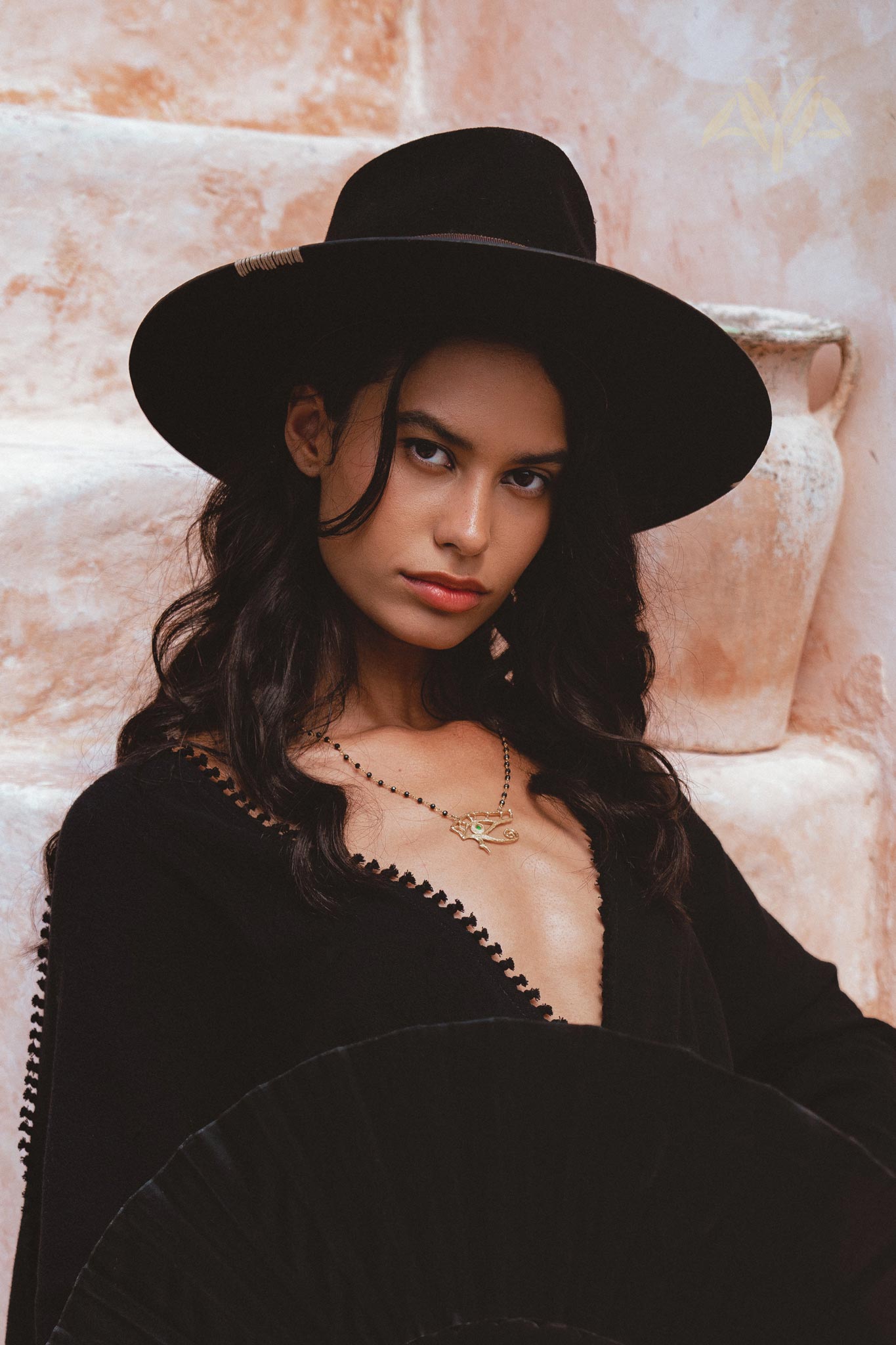 A woman poses confidently in an oversized hand-knotted black jumpsuit, featuring an elegant and asymmetrical design. She wears a stylish black hat and a delicate necklace, set against a warm, textured background that enhances the sophisticated look of the outfit. Perfect for showcasing the contemporary fashion aesthetic of the Oversized Hand Knotted Jumpsuit.