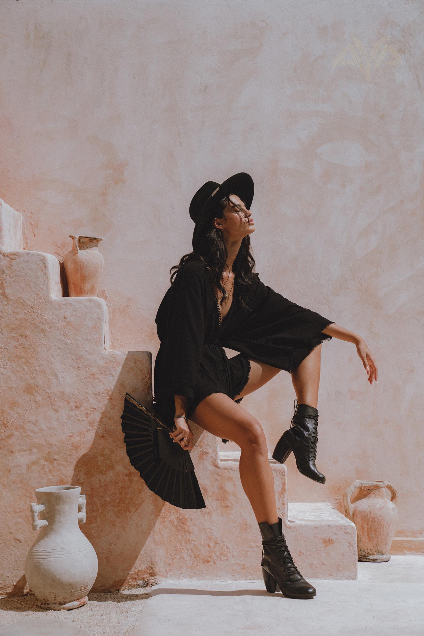 A model poses on a staircase, showcasing an oversized hand-knotted black mini jumpsuit. She wears a wide-brimmed hat and holds a black fan, complemented by stylish black ankle boots. The backdrop features a soft pink wall and a decorative vase, creating an elegant outdoor setting.