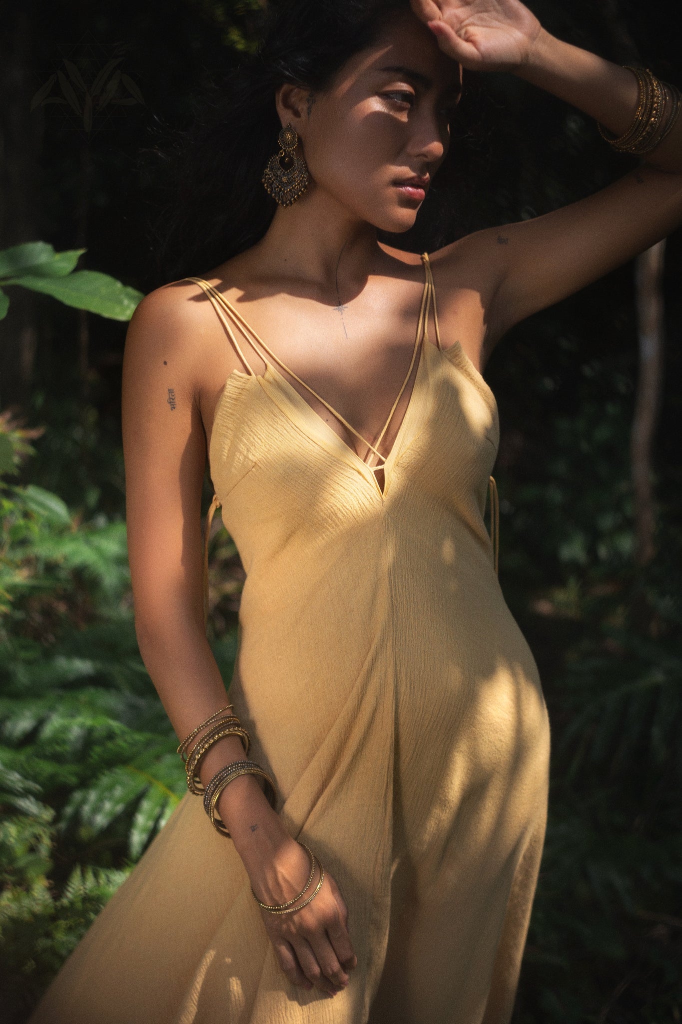 In a sunlit forest, a woman in an AYA Sacred Wear Powder Yellow Padma Dress effortlessly evokes an elegant bohemian wedding moment. Gold earrings and bracelets enhance her look as she stands amidst greenery, her hand shielding her eyes, with lighting that casts a magical glow over the serene scene.