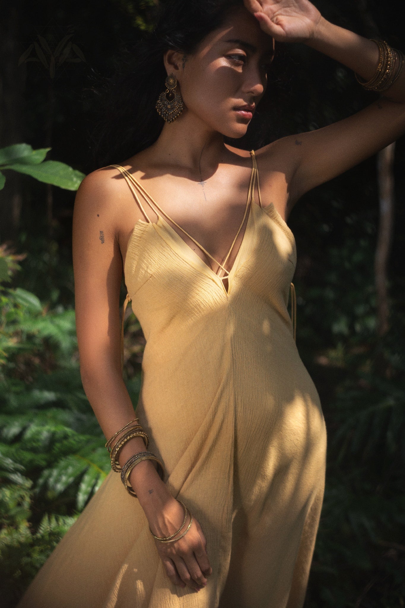 In a sunlit forest, a woman wearing AYA Sacred Wear's Powder Yellow Padma Dress shields her eyes with bejeweled hands. Her intricate earrings glimmer as shadows dance on her face amidst the elegant dress and surrounding foliage.