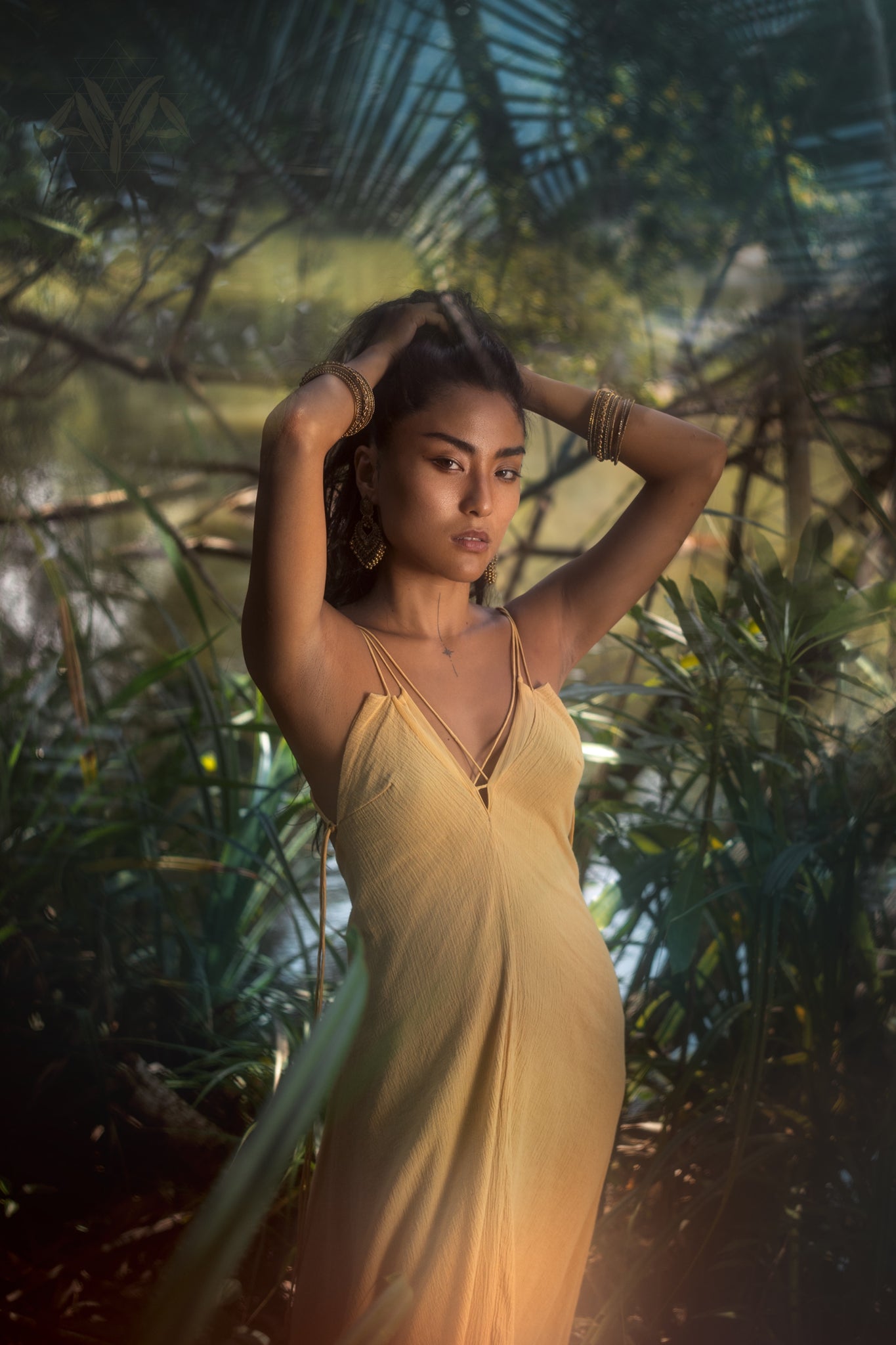 In a lush, sunlit forest, someone wears the Powder Yellow Padma Dress by AYA Sacred Wear. Adorned with bracelets and earrings, their hands in their hair, the shadowed leaves enhance the tranquil atmosphere around this Boho Backless Sleeveless Elegant Dress made of organic handwoven cotton.