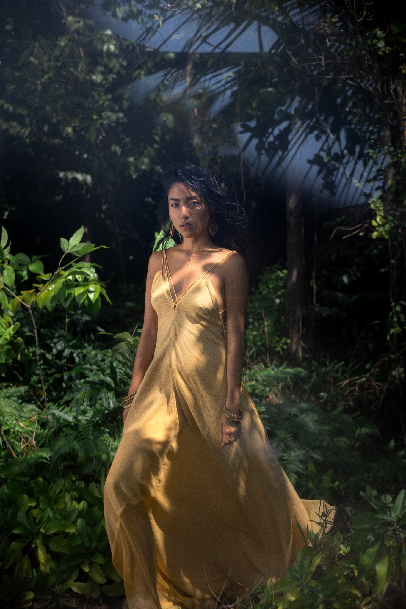 In a lush, green forest, a woman wearing AYA Sacred Wear's Powder Yellow Padma Dress—an elegant, sleeveless boho backless gown—stands serenely. Sunlight filters through the trees, casting shadows as her hair gently flows in the breeze, embodying the essence of this organic handwoven dress.