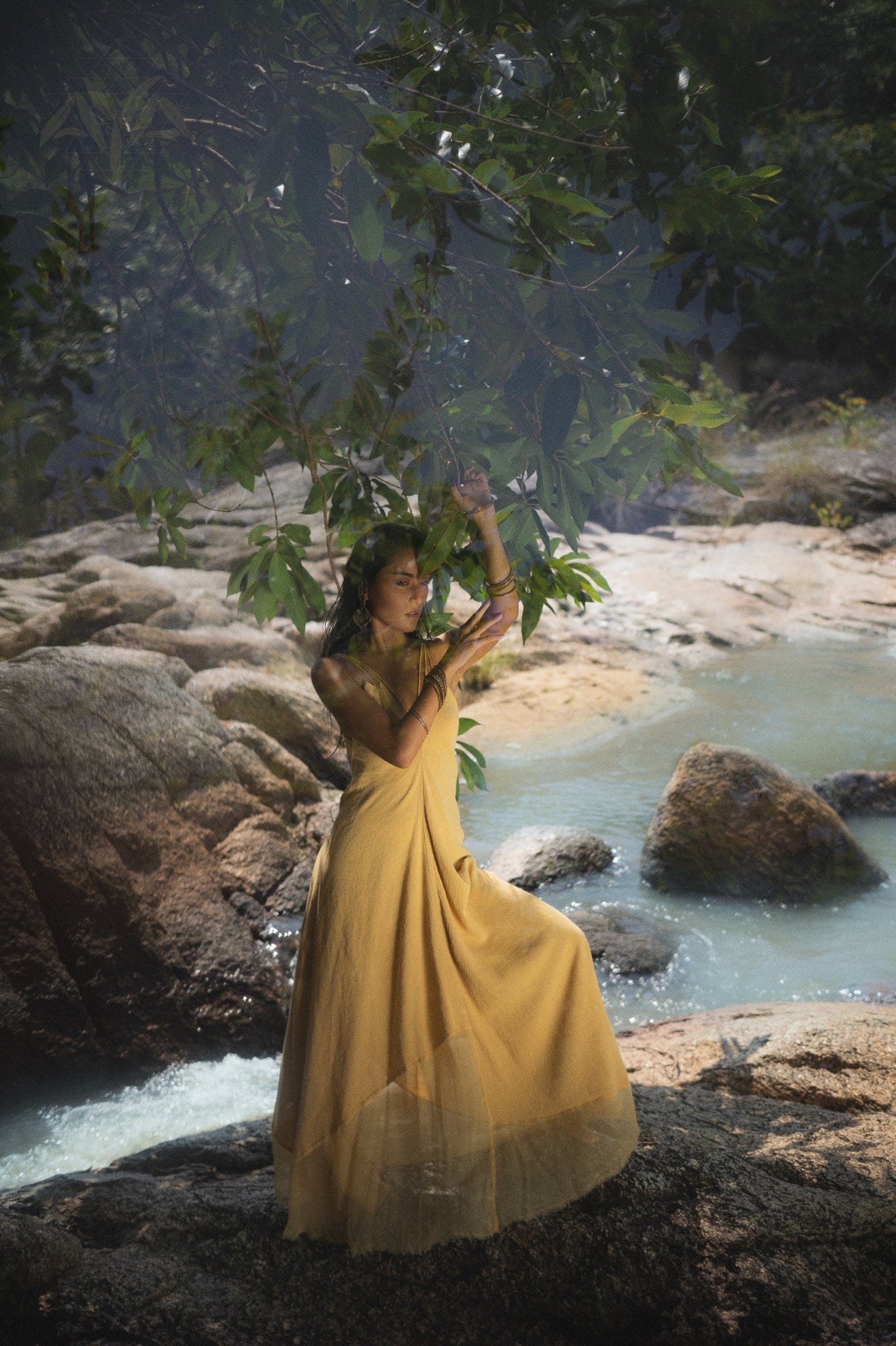 A woman elegantly poses on rocks by a gentle stream in a Powder Yellow Padma Dress from AYA Sacred Wear. She reaches for the branches amid lush greenery, creating a serene bohemian wedding ambiance.